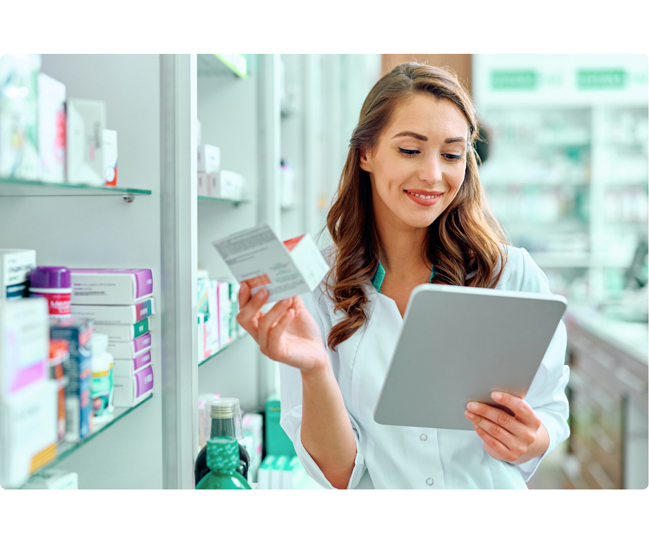 Fundo de uma farmácia, do lado direito há uma mulher branca, com cabelos claros soltos, a mesma está sorrindo, segurando um tablet em uma mão e na outra um medicamento. A mesma está vestindo jaleco branco.